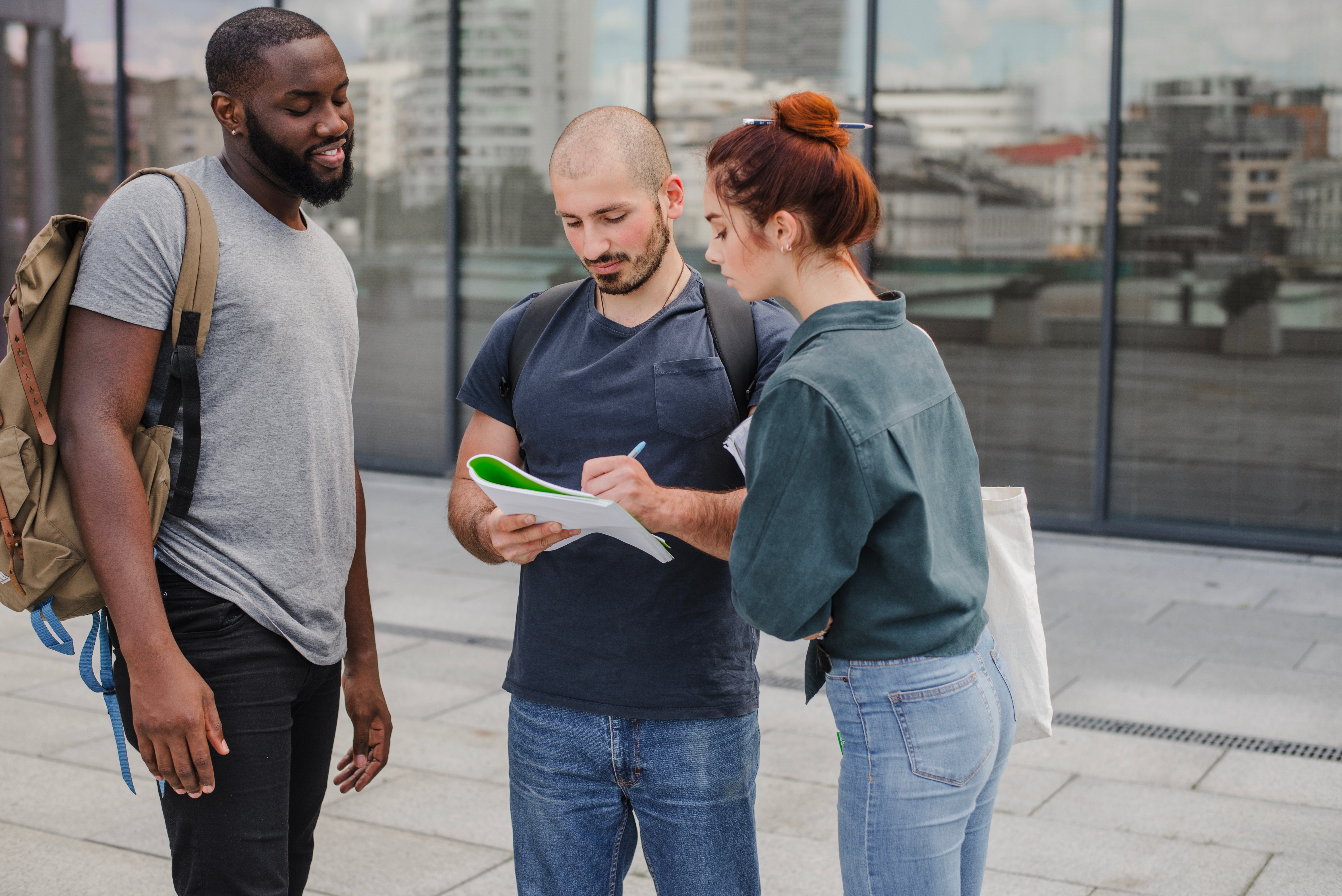 Three students of college age standing outside around a notebook writing notes.